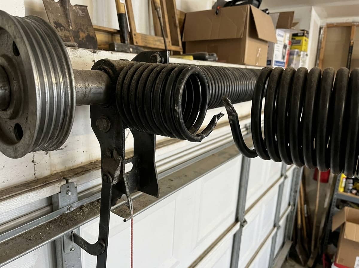 Resorte de torsion negro roto sobre el eje de una puerta seccional cerrada, con un hueco visible donde el acero se partio