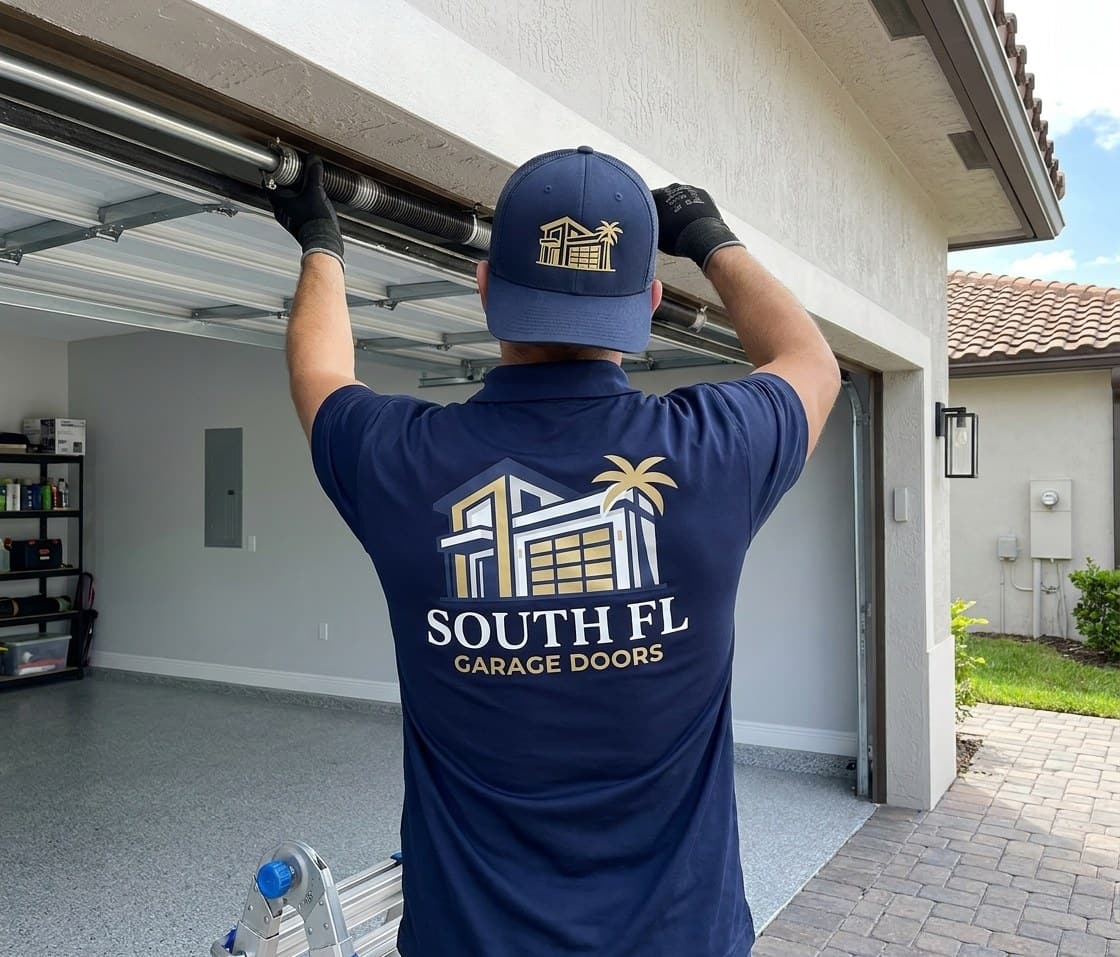 Tecnico con polo y gorra azul marino de South FL Garage Doors, visto de espaldas sobre una escalera, con ambas manos enguantadas en el eje de resortes sobre una puerta de garaje abierta